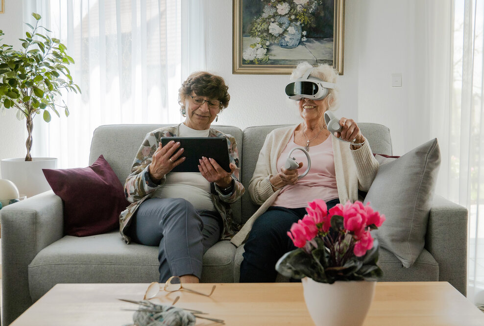 Seniorinnen mit VR-Brille und Tablet auf einem hellgrauen Sofa bei der Anwendung von teora mind
