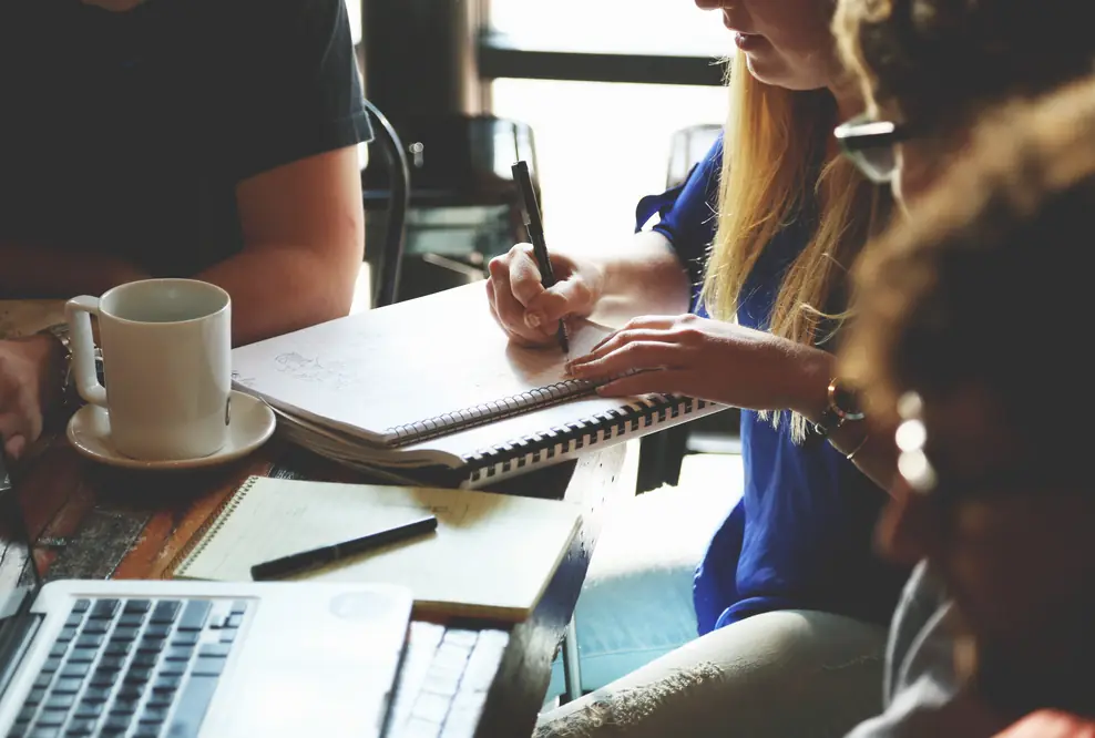 Gründerteam bei der gemeinsamen Arbeit am Tisch – Notizbücher, Laptop und Kaffeetasse im Einsatz, während sie einen Businessplan erstellen und Strategien für ihre Unternehmensgründung entwickeln..