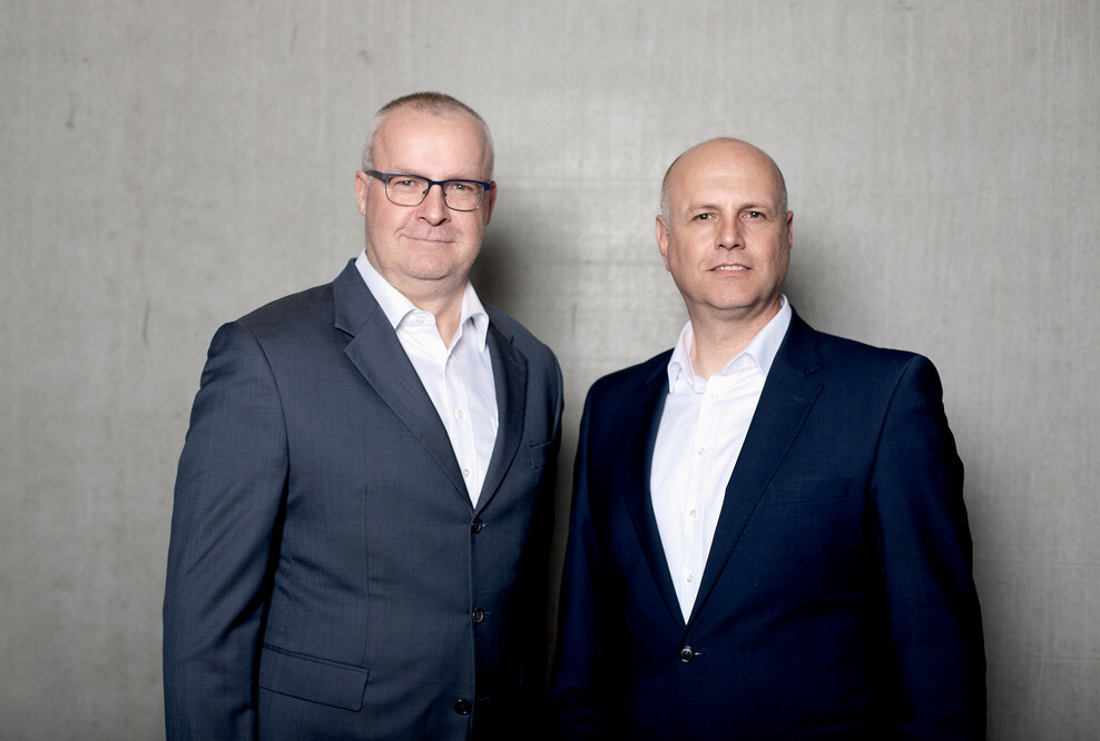 Die Geschäftsführer von Persus Personal, Martin Bach und Andreas Nusko stehen in Anzug mit weißem Hemd vor einer Betonwand
