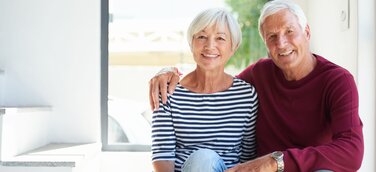 Lächelndes Seniorenpaar sitzt im Tageslicht vor einem Fenster seines Eigenheims nach Immobilien Teilverkauf.