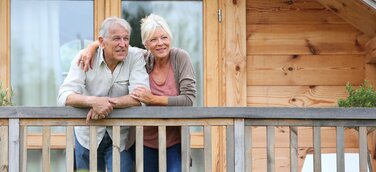 Älteres Paar steht lächelnd auf Balkon mit Holzfassade im Hintergrund.
