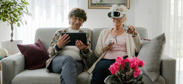 Seniorinnen mit VR-Brille und Tablet auf einem hellgrauen Sofa bei der Anwendung von teora mind
