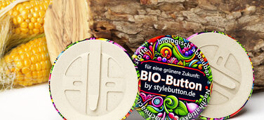 3 biologisch abbaubare Buttons von stylebutton.de, im Hintergrund sind ein Holzscheit und Maiskolben zu sehen