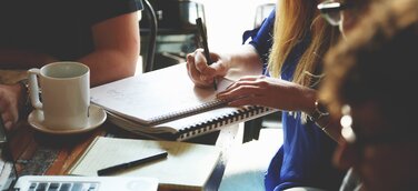 Gründerteam bei der gemeinsamen Arbeit am Tisch – Notizbücher, Laptop und Kaffeetasse im Einsatz, während sie einen Businessplan erstellen und Strategien für ihre Unternehmensgründung entwickeln..