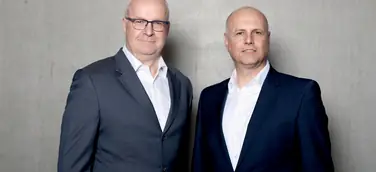 Die Geschäftsführer von Persus Personal, Martin Bach und Andreas Nusko stehen in Anzug mit weißem Hemd vor einer Betonwand