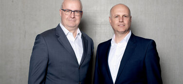 Die Geschäftsführer von Persus Personal, Martin Bach und Andreas Nusko stehen in Anzug mit weißem Hemd vor einer Betonwand