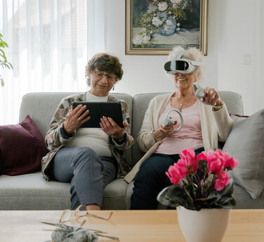 Seniorinnen mit VR-Brille und Tablet auf einem hellgrauen Sofa bei der Anwendung von teora mind