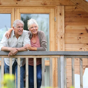 Älteres Paar steht lächelnd auf Balkon mit Holzfassade im Hintergrund.