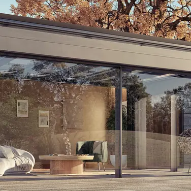 Modernes Terrassendach aus Glas und Aluminium, harmonisch an ein Einfamilienhaus mit Natursteinfassade angebaut. Die große Glasfront öffnet den Wohnraum zum Garten, umgeben von herbstlicher Natur und warmem Licht. Ein stilvoller Lounge-Bereich mit Sofa und Couchtisch lädt zum Entspannen ein – Symbol für modernes Wohnen und #Draußenzuhause mit Sachsen Terrassen & Glas.