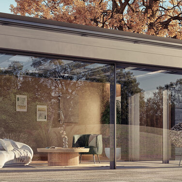 Modernes Terrassendach aus Glas und Aluminium, harmonisch an ein Einfamilienhaus mit Natursteinfassade angebaut. Die große Glasfront öffnet den Wohnraum zum Garten, umgeben von herbstlicher Natur und warmem Licht. Ein stilvoller Lounge-Bereich mit Sofa und Couchtisch lädt zum Entspannen ein – Symbol für modernes Wohnen und #Draußenzuhause mit Sachsen Terrassen & Glas.