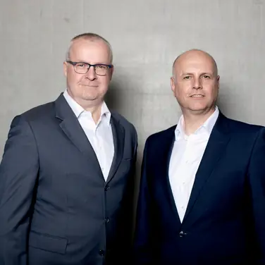 Die Geschäftsführer von Persus Personal, Martin Bach und Andreas Nusko stehen in Anzug mit weißem Hemd vor einer Betonwand