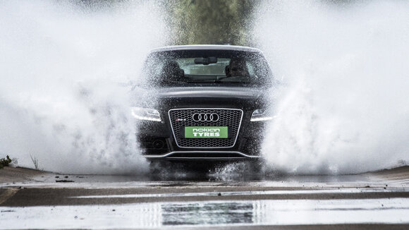 Auto Frontansicht mit Nummernschild Nokian Tyres fährt auf nasser Fahrbahn, Gischt spritzt sehr hoch links und rechts am Auto