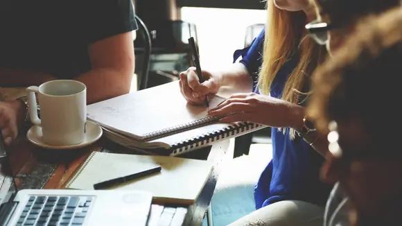 Gründerteam bei der gemeinsamen Arbeit am Tisch – Notizbücher, Laptop und Kaffeetasse im Einsatz, während sie einen Businessplan erstellen und Strategien für ihre Unternehmensgründung entwickeln..