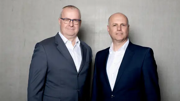 Die Geschäftsführer von Persus Personal, Martin Bach und Andreas Nusko stehen in Anzug mit weißem Hemd vor einer Betonwand