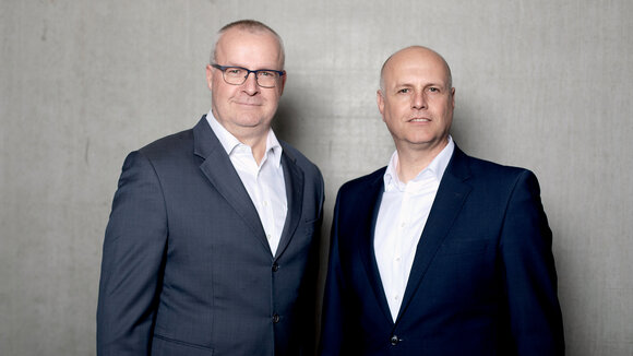 Die Geschäftsführer von Persus Personal, Martin Bach und Andreas Nusko stehen in Anzug mit weißem Hemd vor einer Betonwand