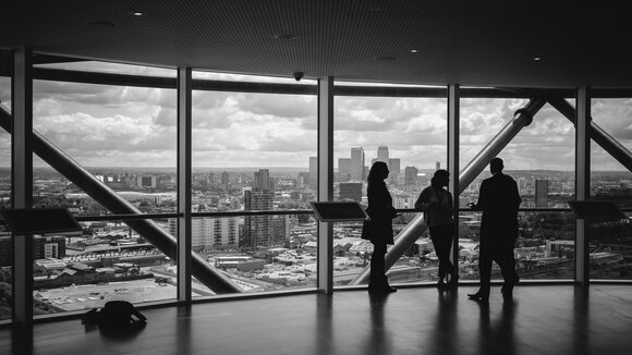 Drei Geschäftsleute stehen als Silhouetten vor einer Panoramafensterfront in einem modernen Hochhaus und besprechen ihre Marketingstrategie — symbolisch für strategische Unternehmensberatung im Bereich KMU Marketing.